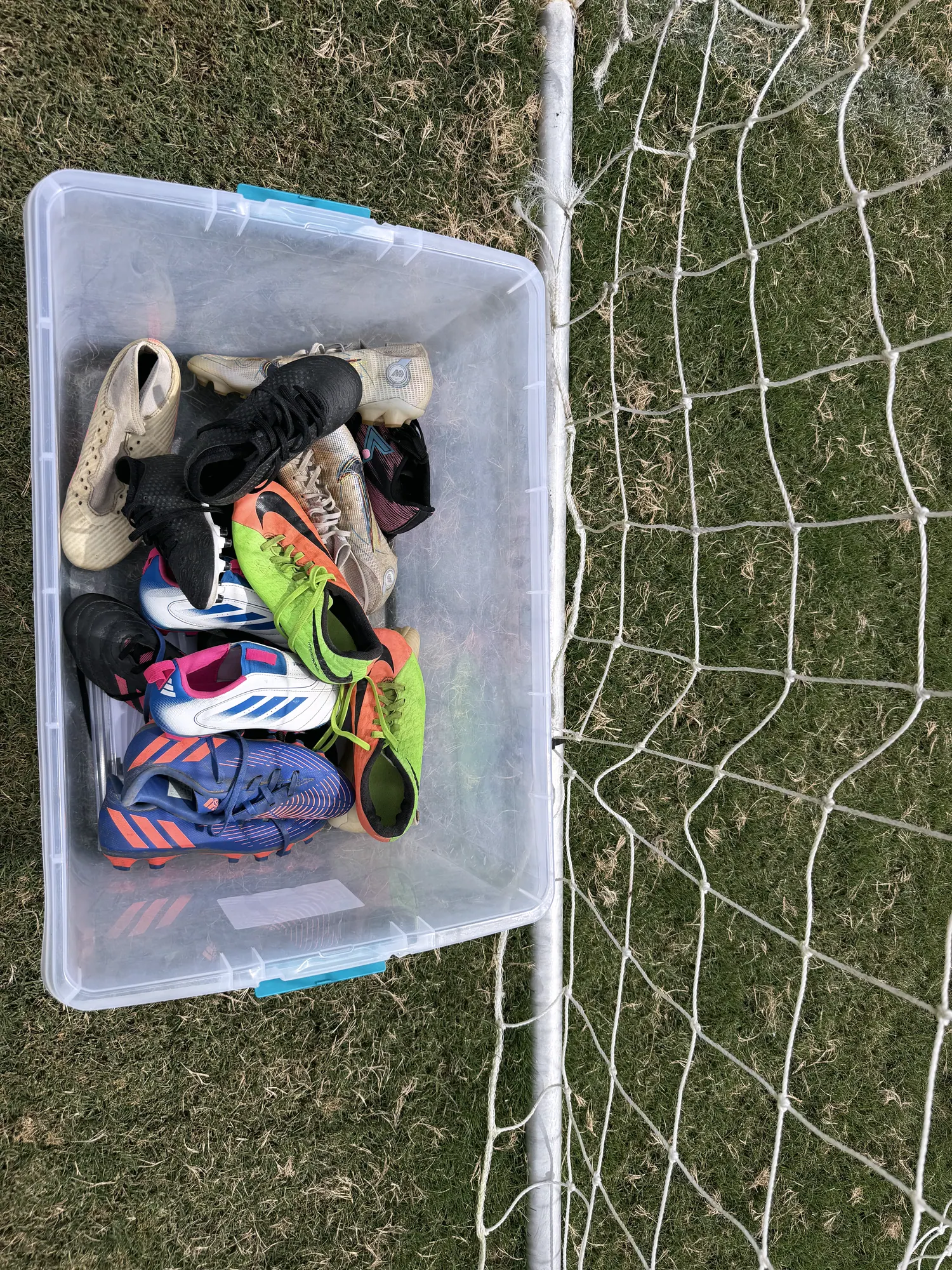 A clear bin filled with donated cleats of every colour and size, propped against a goal net