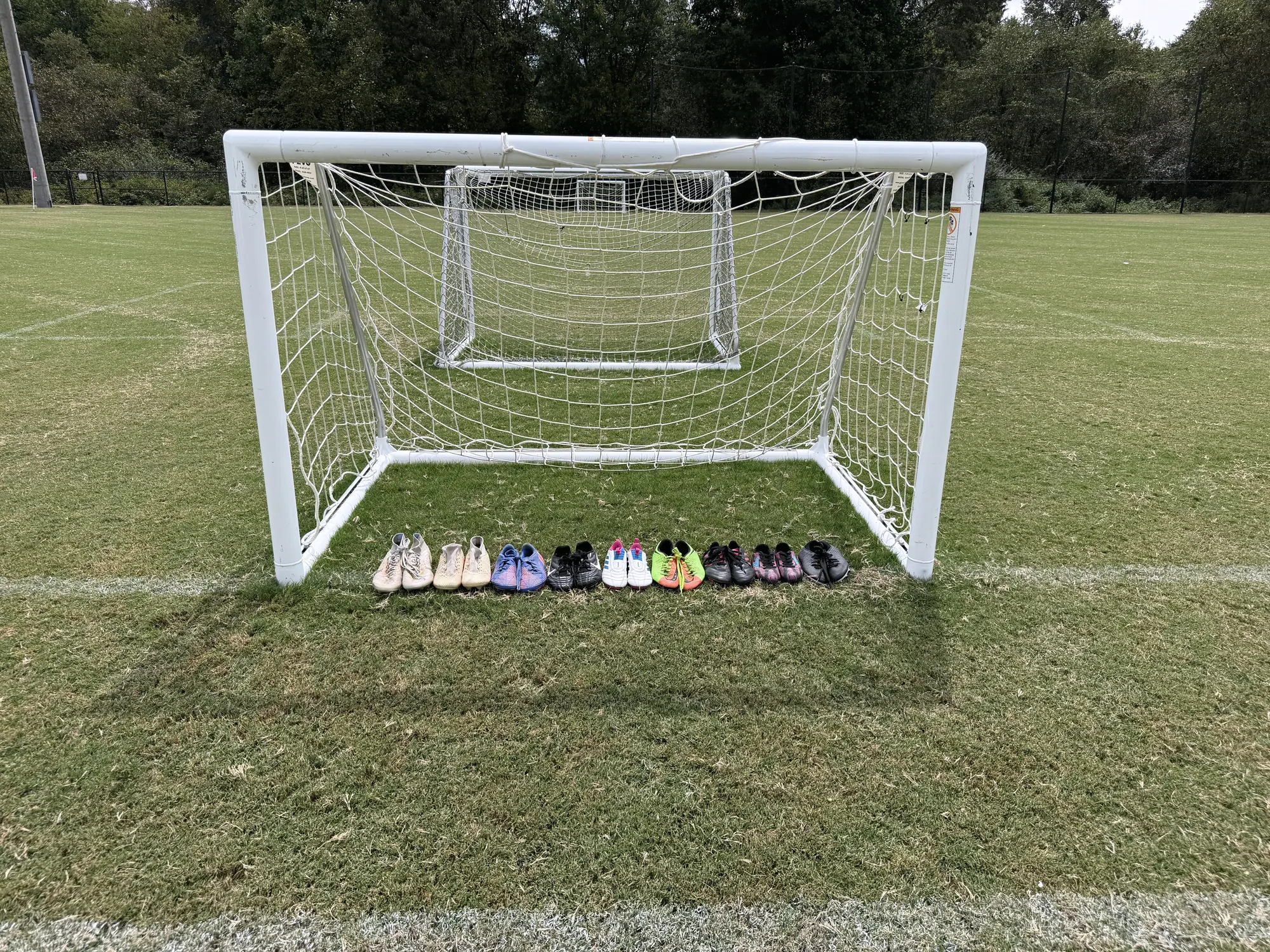 A row of donated soccer cleats lined up in pairs across the mouth of an empty goal on a grass field